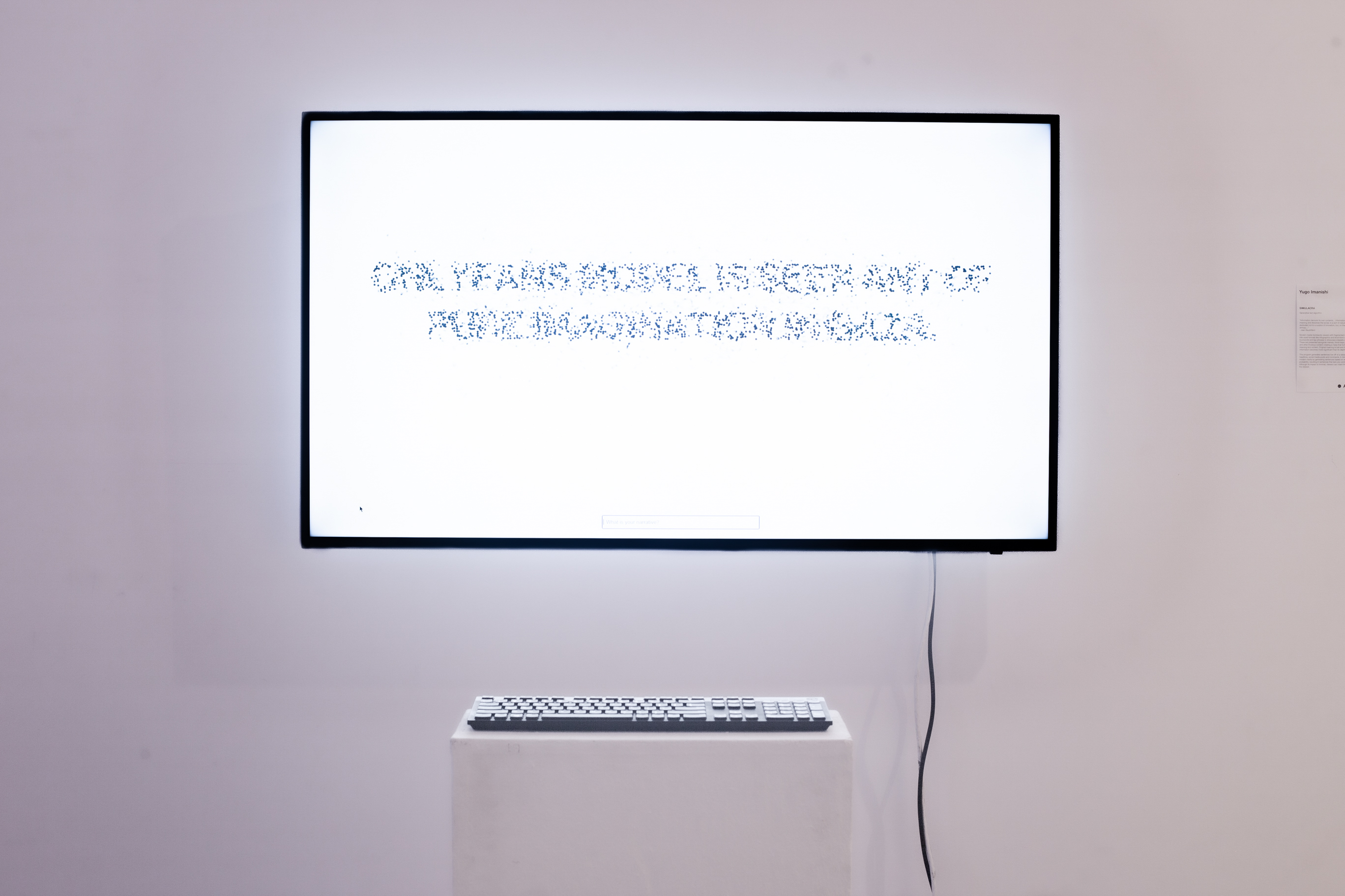 A TV placed on a white wall with a podium in front of it with a keyboard. The TV has a white picture with incomprehensive text drawn by dots.